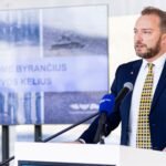 Kostiumu vilkintis vyras kalba prie tribūnos su mikrofonais