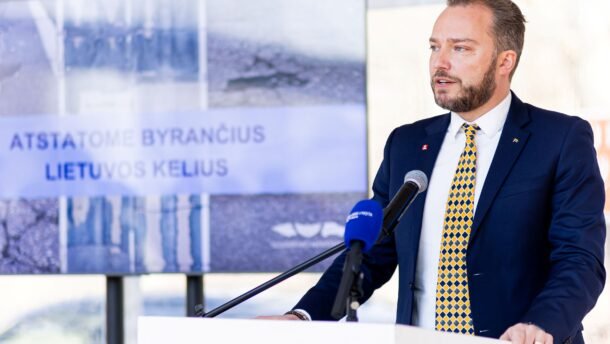 Kostiumu vilkintis vyras kalba prie tribūnos su mikrofonais
