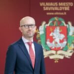 Kostiumu vilkintis vyras stovi priešais Vilniaus miesto savivaldybės herbą