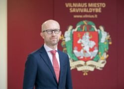 Kostiumu vilkintis vyras stovi priešais Vilniaus miesto savivaldybės herbą