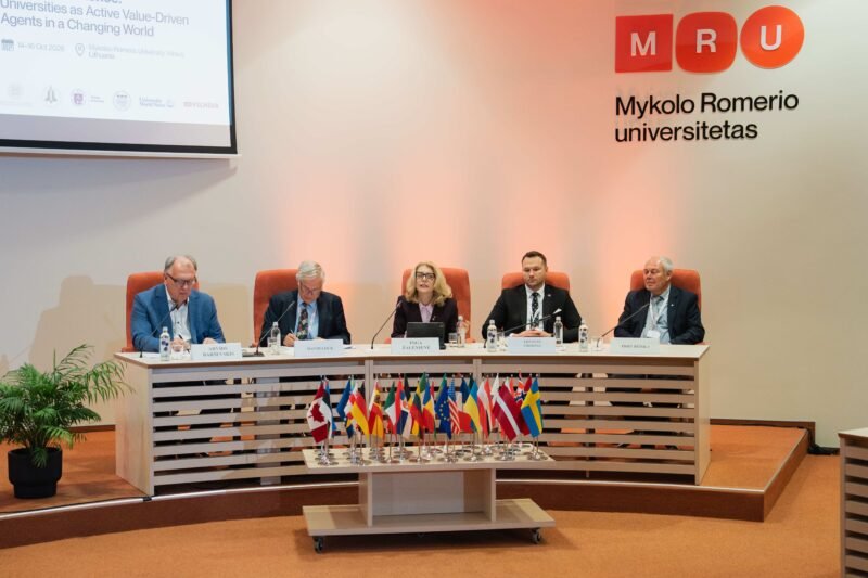 Penki universitetų atstovai sėdi prie konferencijų stalo su įvairių šalių vėliavėlėmis