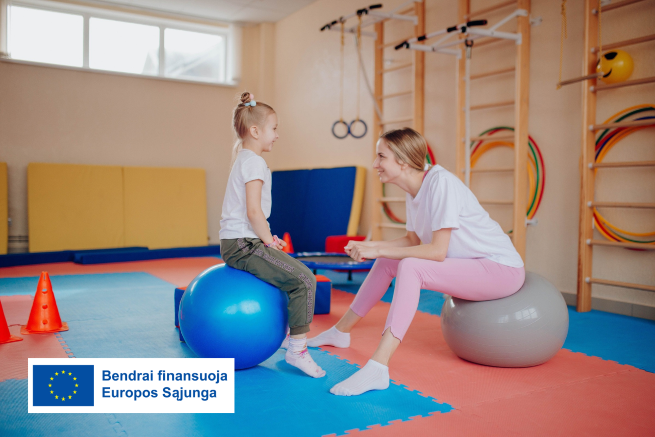 Kineziterapeutė su mergaite sėdi ant gimnastikos kamuolių vaikų mankštos salėje