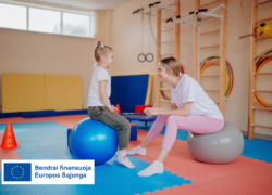Kineziterapeutė su mergaite sėdi ant gimnastikos kamuolių vaikų mankštos salėje