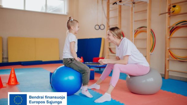 Kineziterapeutė su mergaite sėdi ant gimnastikos kamuolių vaikų mankštos salėje