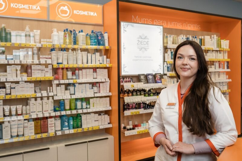 Vaistininkė balta uniforma stovi kosmetikos prekių skyriuje prie lentynų