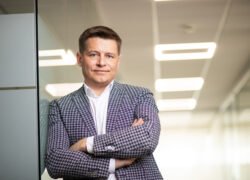 Verslininkas sukryžiuotomis rankomis stovi šviesiame biuro koridoriuje