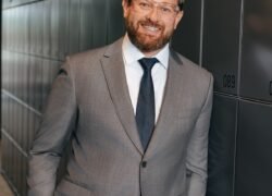 Besirūsmantis vyras su pilku kostiumu stovi prie tamsių spintelių