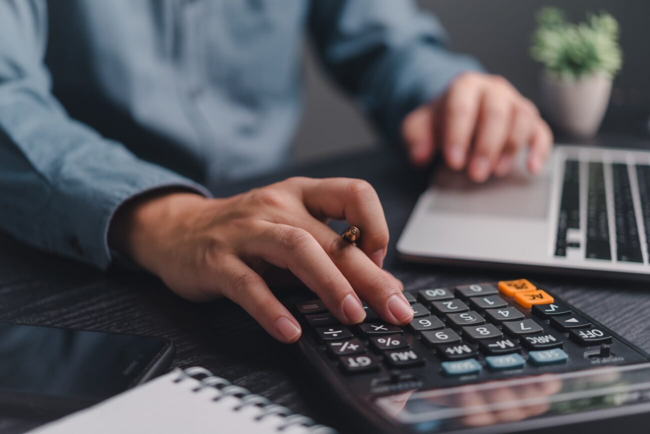 Verslininkas skaičiuoja finansus kalkuliatoriumi prie nešiojamojo kompiuterio