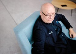 Plikagalvis vyras su akiniais sėdi šviesiai mėlyname fotelyje