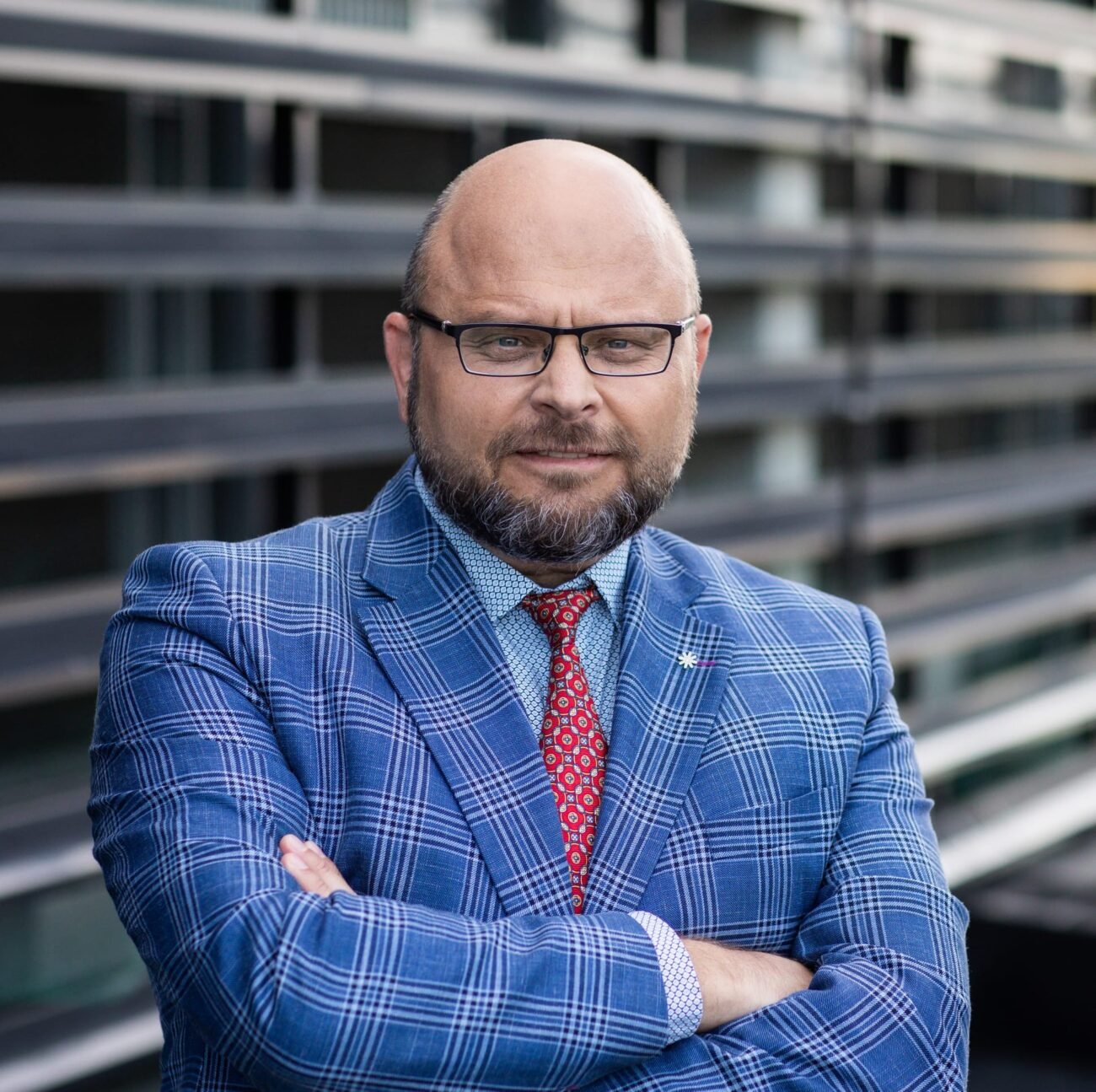 Bardą ir akinius dėvintis vyras mėlynu languotu kostiumu sukryžiavęs rankas