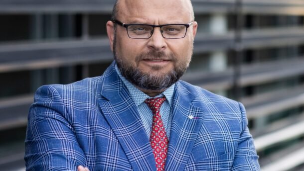 Bardą ir akinius dėvintis vyras mėlynu languotu kostiumu sukryžiavęs rankas