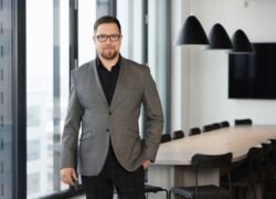 Vyras su akiniais ir švarku stovi moderniame biure prie konferencijų stalo