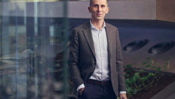 Vyras su švarku stovi modernioje biuro erdvėje prie stiklinių sienų