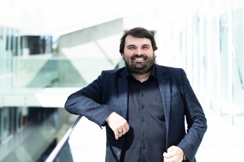 Barzdotas vyras tamsiu švarku stovi šviesiame moderniame pastate