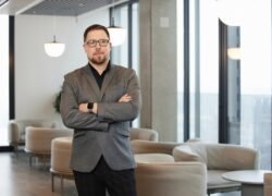 Vyras su akiniais ir sukryžiuotomis rankomis stovi moderniame biuro interjere