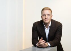 Prie stalo sėdintis vyras su tamsiu švarku ir baltais marškiniais