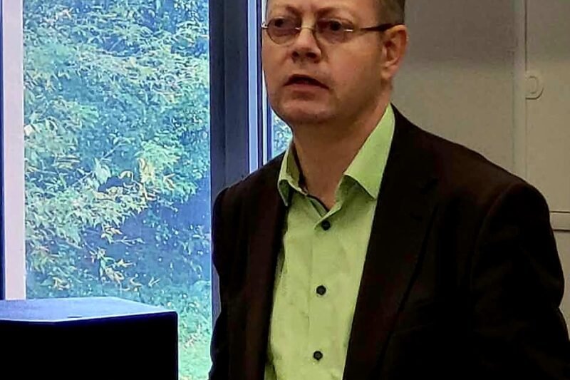 Akiniais pasipuošęs vyras stovi prie medinės tribūnos šalia kompiuterio