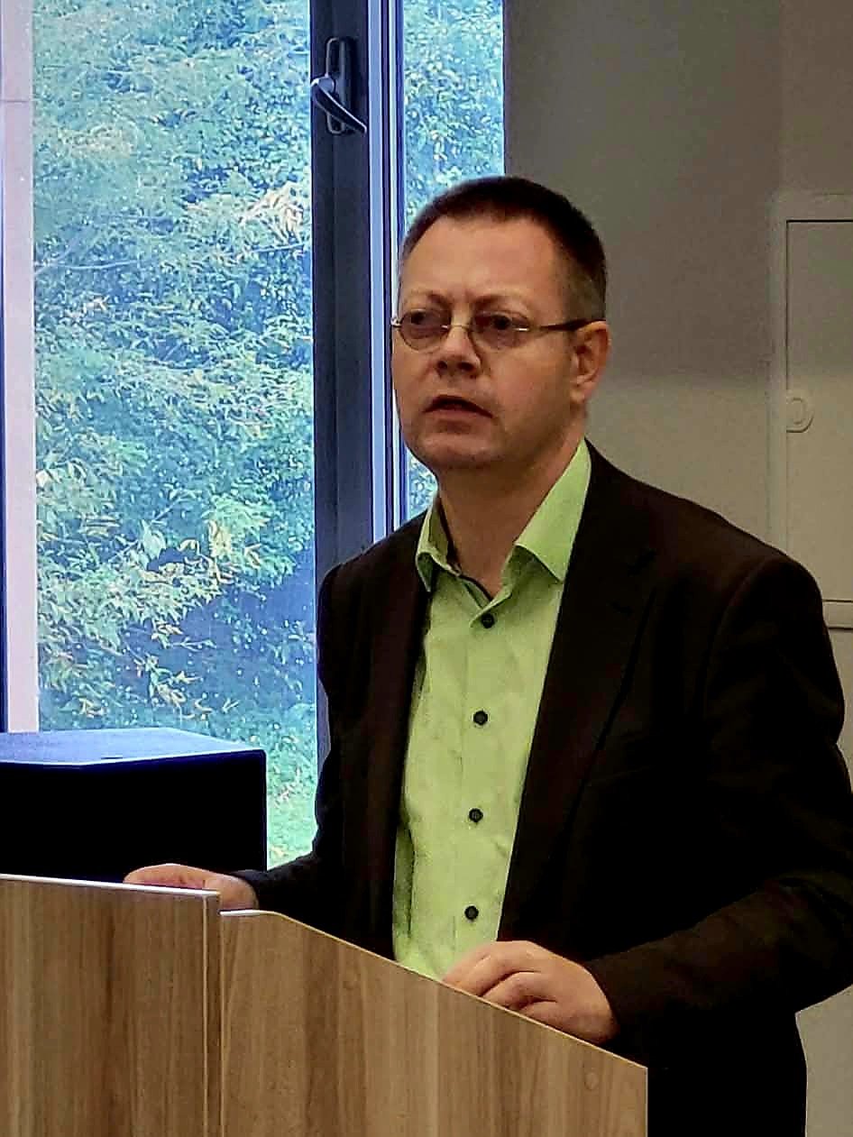 Akiniais pasipuošęs vyras stovi prie medinės tribūnos šalia kompiuterio