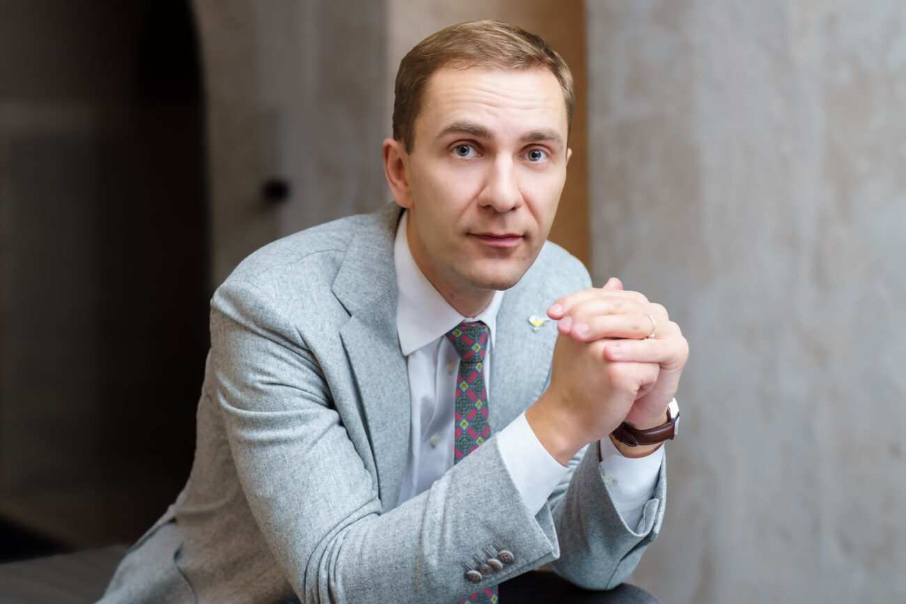 Vyras su šviesiai pilku kostiumu sėdi sukabinęs rankas