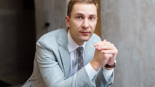 Vyras su šviesiai pilku kostiumu sėdi sukabinęs rankas