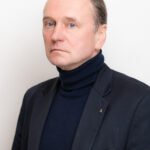 Rimto žvilgsnio vyras tamsiu švarku ir aukštu kaklu megztiniu