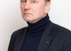 Rimto žvilgsnio vyras tamsiu švarku ir aukštu kaklu megztiniu