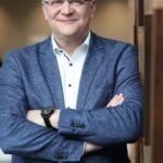 Besišypsantis žilas vyras su akiniais ir sukryžiuotomis rankomis mėlyname švarke