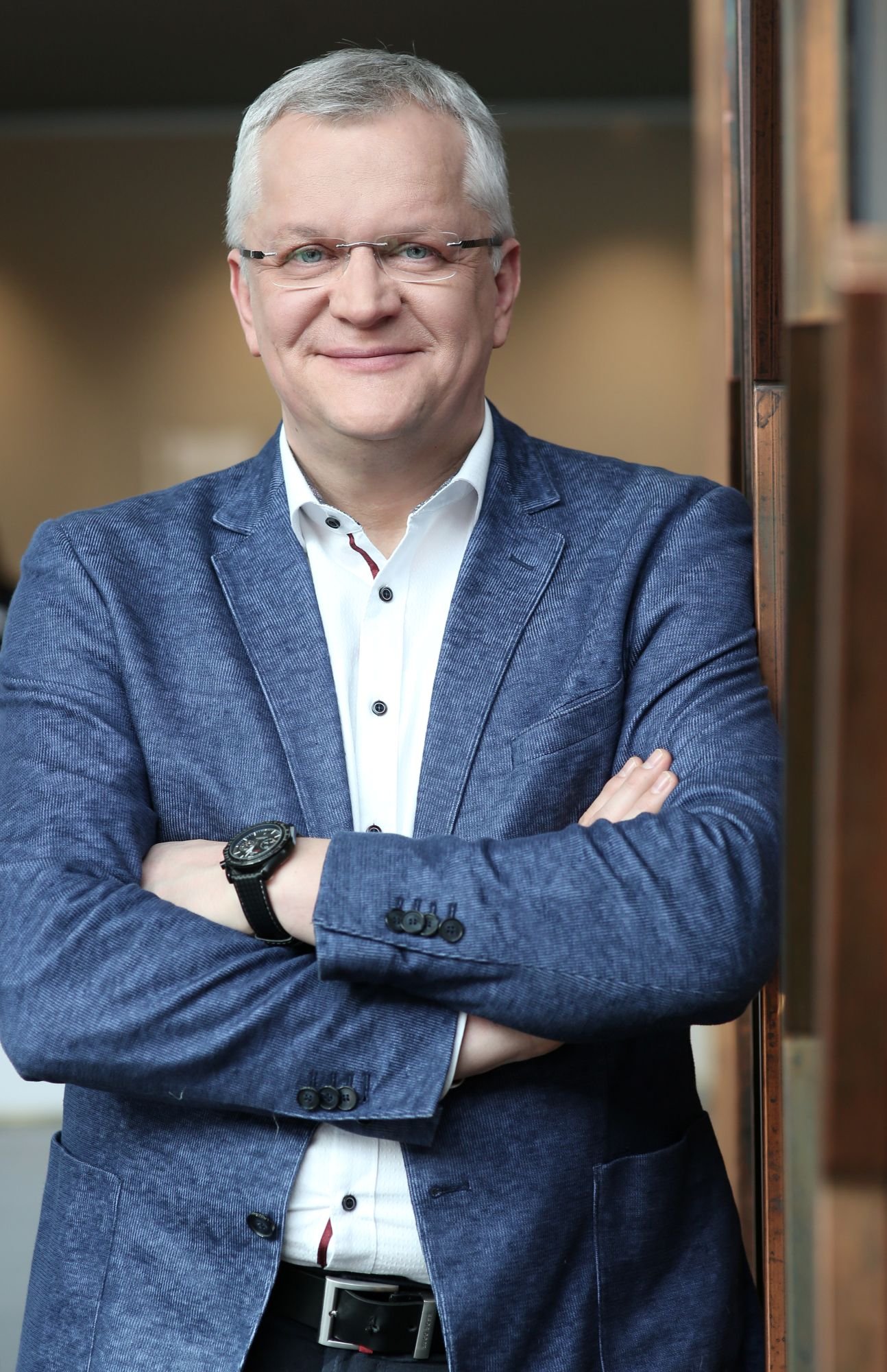Besišypsantis žilas vyras su akiniais ir sukryžiuotomis rankomis mėlyname švarke