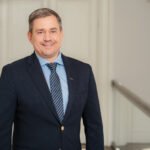 Besijuokiantis vyras tamsiu kostiumu stovi prie laiptų