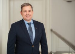 Besijuokiantis vyras tamsiu kostiumu stovi prie laiptų