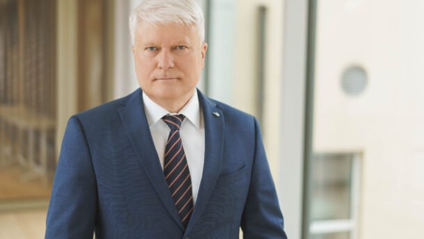 Baltaplaukis vyras mėlynu kostiumu pozuoja prie pastato