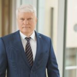 Žilas vyras su mėlynu kostiumu stovi prie pastato