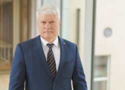 Žilas vyras su mėlynu kostiumu stovi prie pastato
